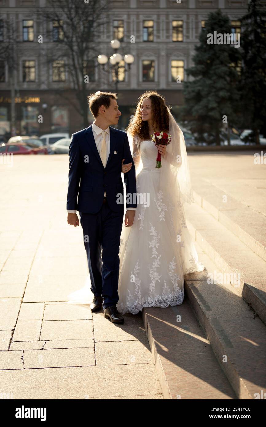 Wunderschönes, gerade verheiratetes Paar, das an sonnigem Tag auf der Straße spaziert Stockfoto