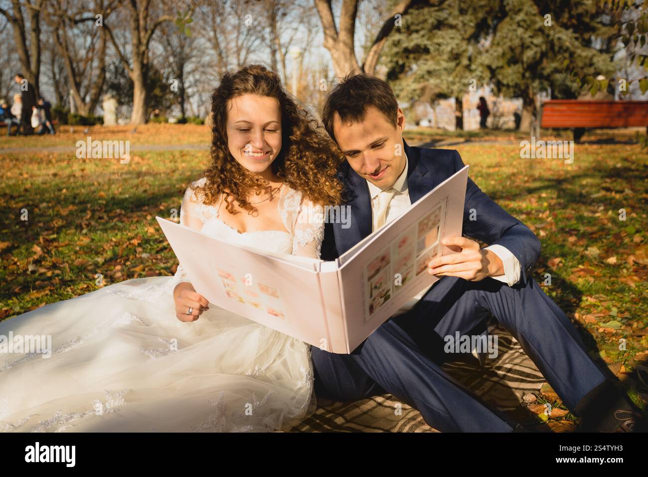 Wunderschönes, gerade verheiratetes Paar, das im Park auf Gras sitzt und Fotos ansieht Stockfoto