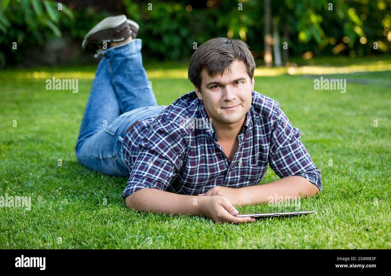 Junger lächelnder Mann liegend mit Tablet auf Rasen im park Stockfoto