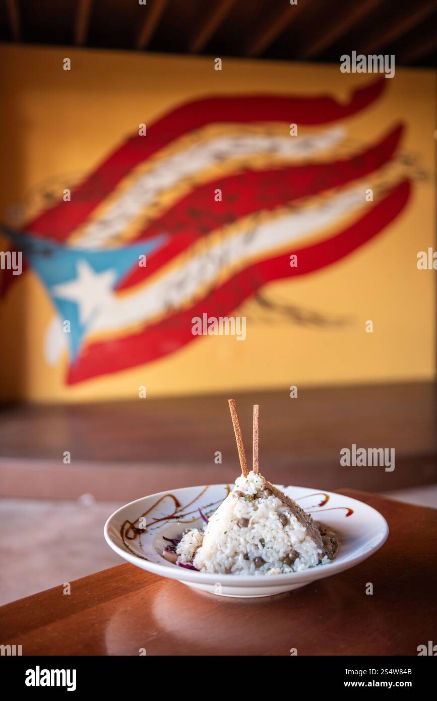 Puerto-ricanischer Arroz con Gandules (Reis und Taubenerbsen) kunstvoll zu einer Pyramide geformt auf einem garnierten Teller mit Puerto-ricanischer Flagge Wandgemälde in bac Stockfoto