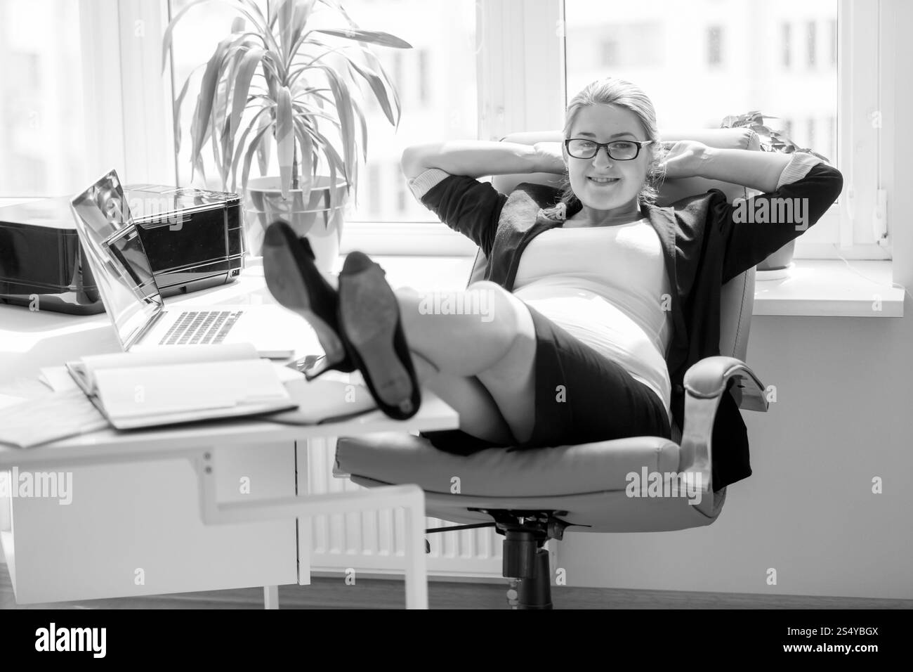 Schwarz-weiß-Bild einer lächelnden schwangeren Geschäftsfrau, die auf einem Stuhl sitzt und Beine auf einem Bürotisch hält Stockfoto