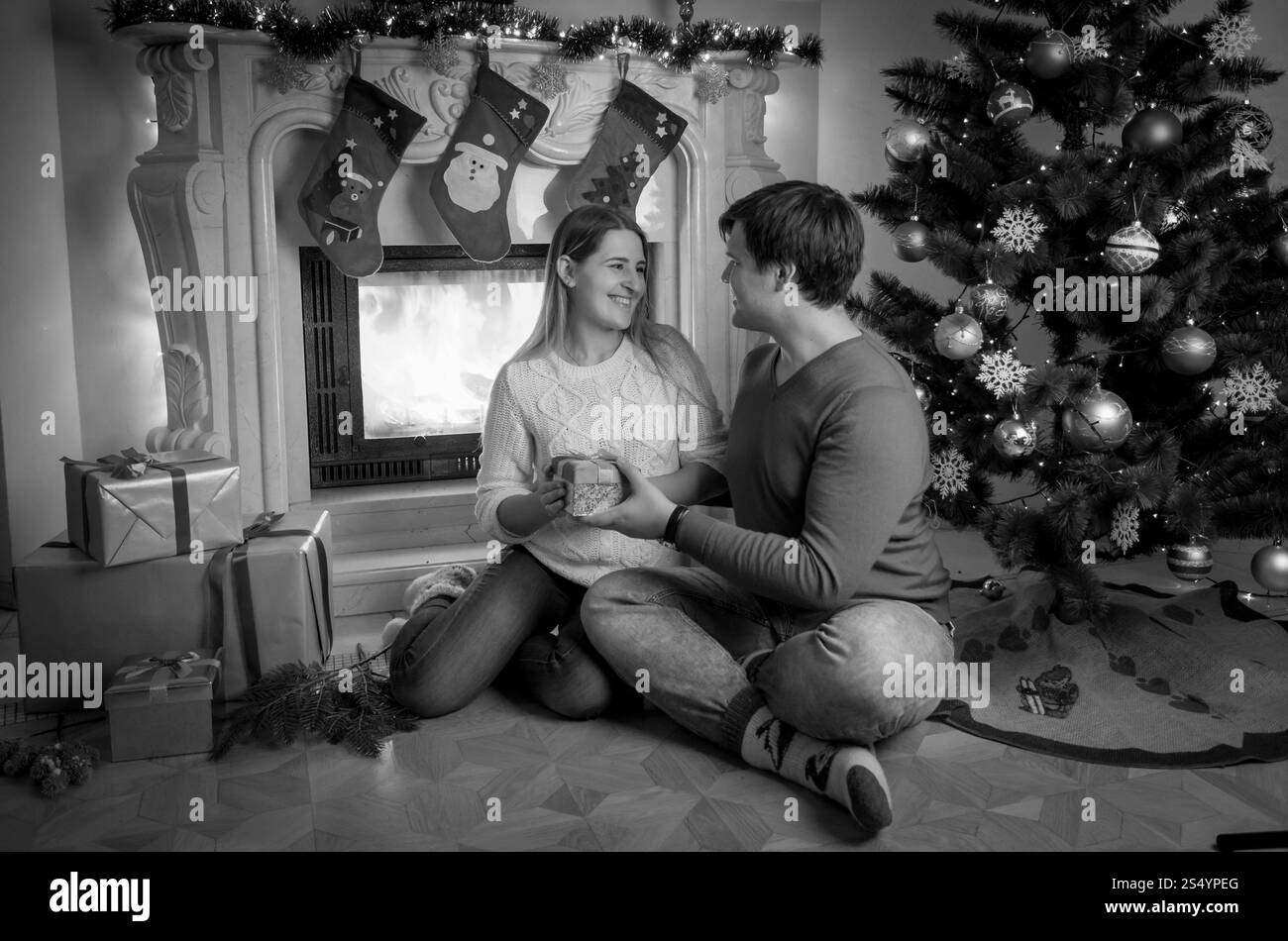 Schwarz-weiß-Bild eines hübschen jungen Mannes, der mit Frau am Kamin sitzt und ihr Weihnachtsgeschenk gibt Stockfoto