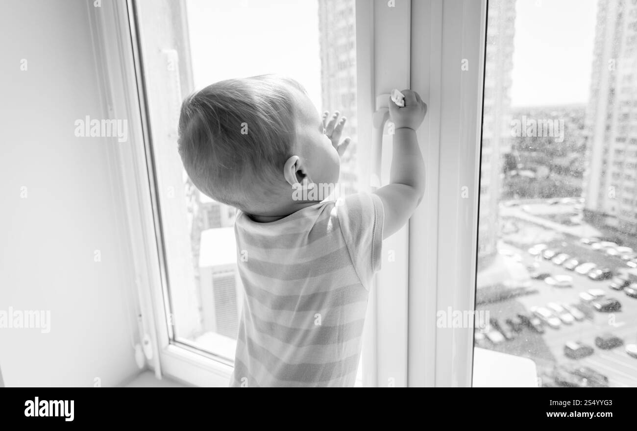 Schwarzweißbild eines Babys in Gefahr. Der Junge zieht am Griff des Fensters Stockfoto