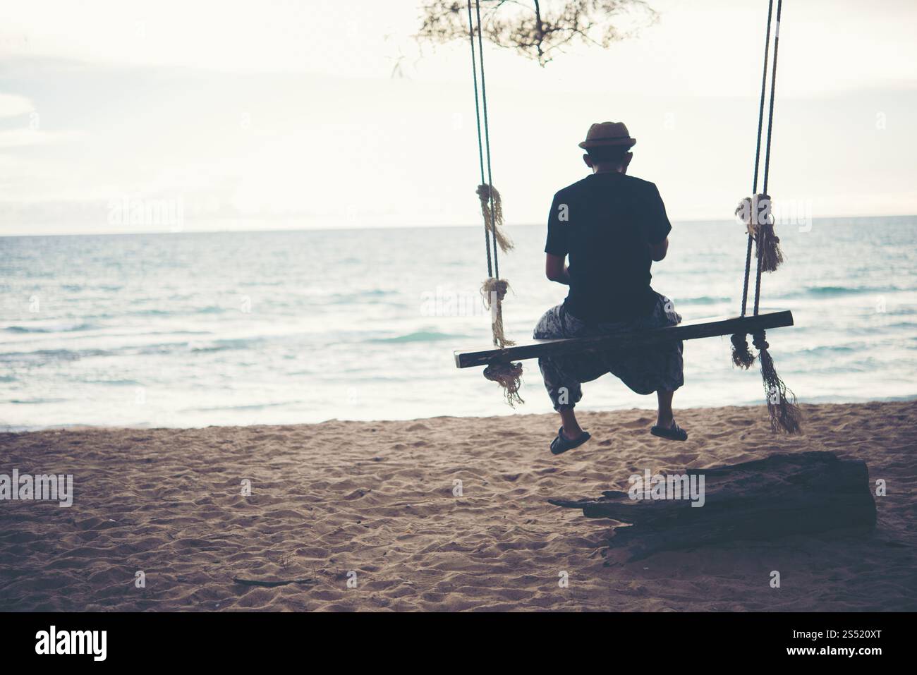 Junger Mann, der den Sonnenuntergang alleine beobachtet, auf Schaukeln am Strand sitzt. Junger Mann, der den Sonnenuntergang alleine beobachtet und am Strand bei Sonnenuntergang auf Schaukeln sitzt Stockfoto