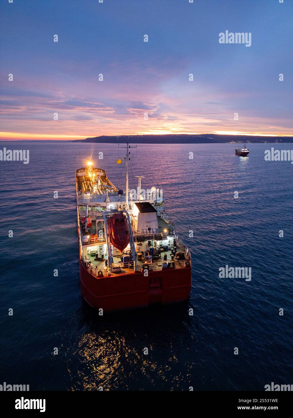 Aus der Vogelperspektive eines hell beleuchteten Tankschiffes auf dem Meer während der Dämmerung, umgeben von offenem Wasser und einem leuchtenden Horizont Stockfoto