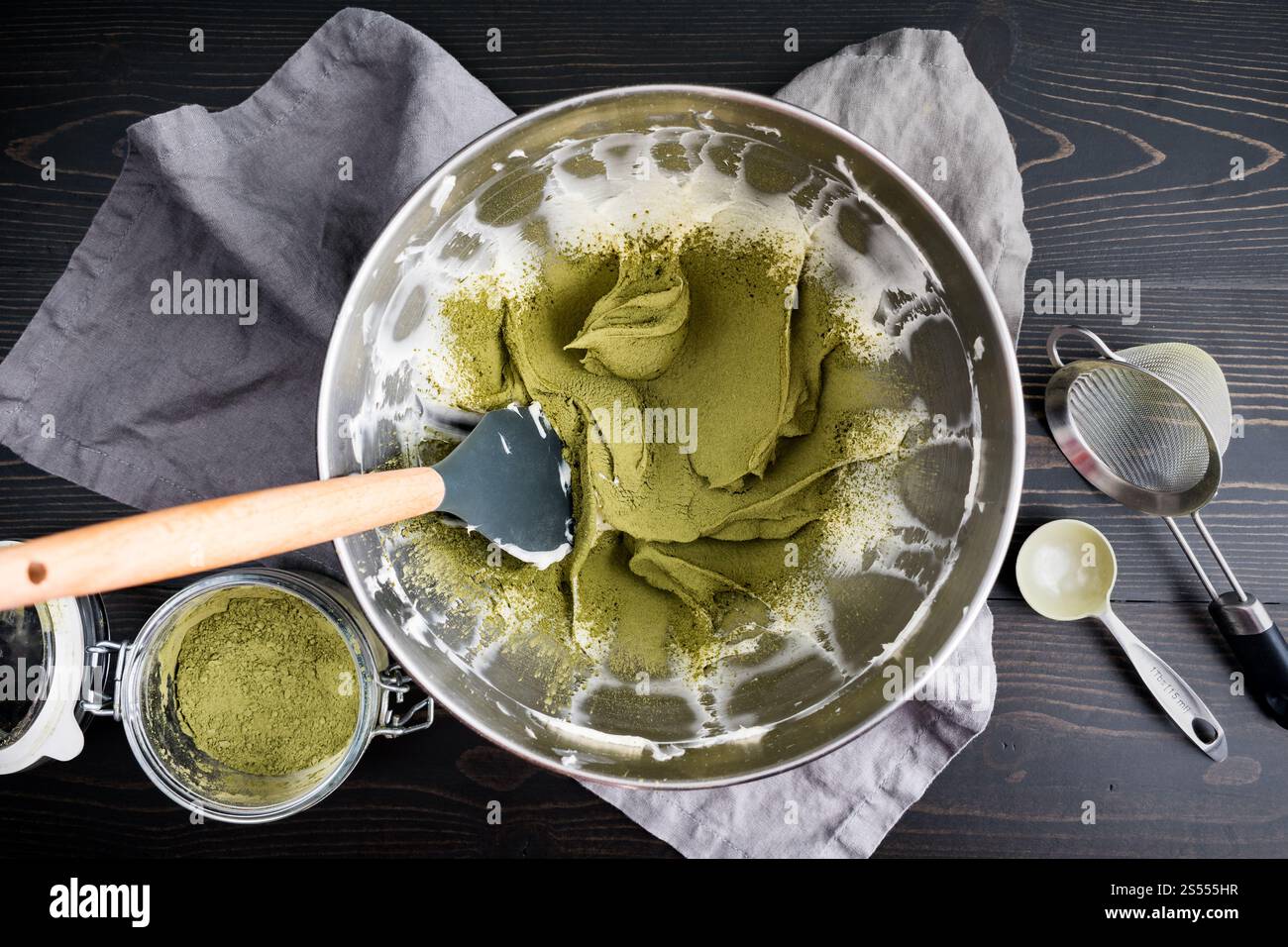 Matcha über Schlagsahne gestreut: Grünes Teepulver, das in einer Metallschüssel mit Spachtel zu verschlagenem Frischkäse gesiebt wird Stockfoto