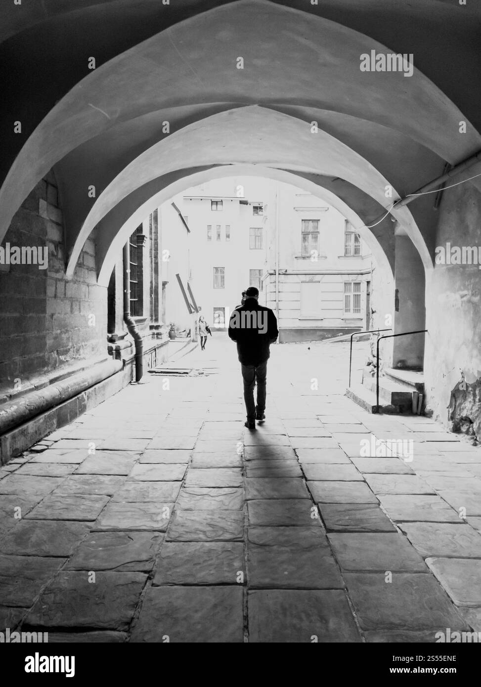 Schwarz-weiß-Foto einer männlichen Silhouette, die auf einer alten schmalen Straße unter einem großen Bogen läuft. Schwarz-weiß-Bild einer männlichen Silhouette, die auf altem schmalen Fuß geht Stockfoto