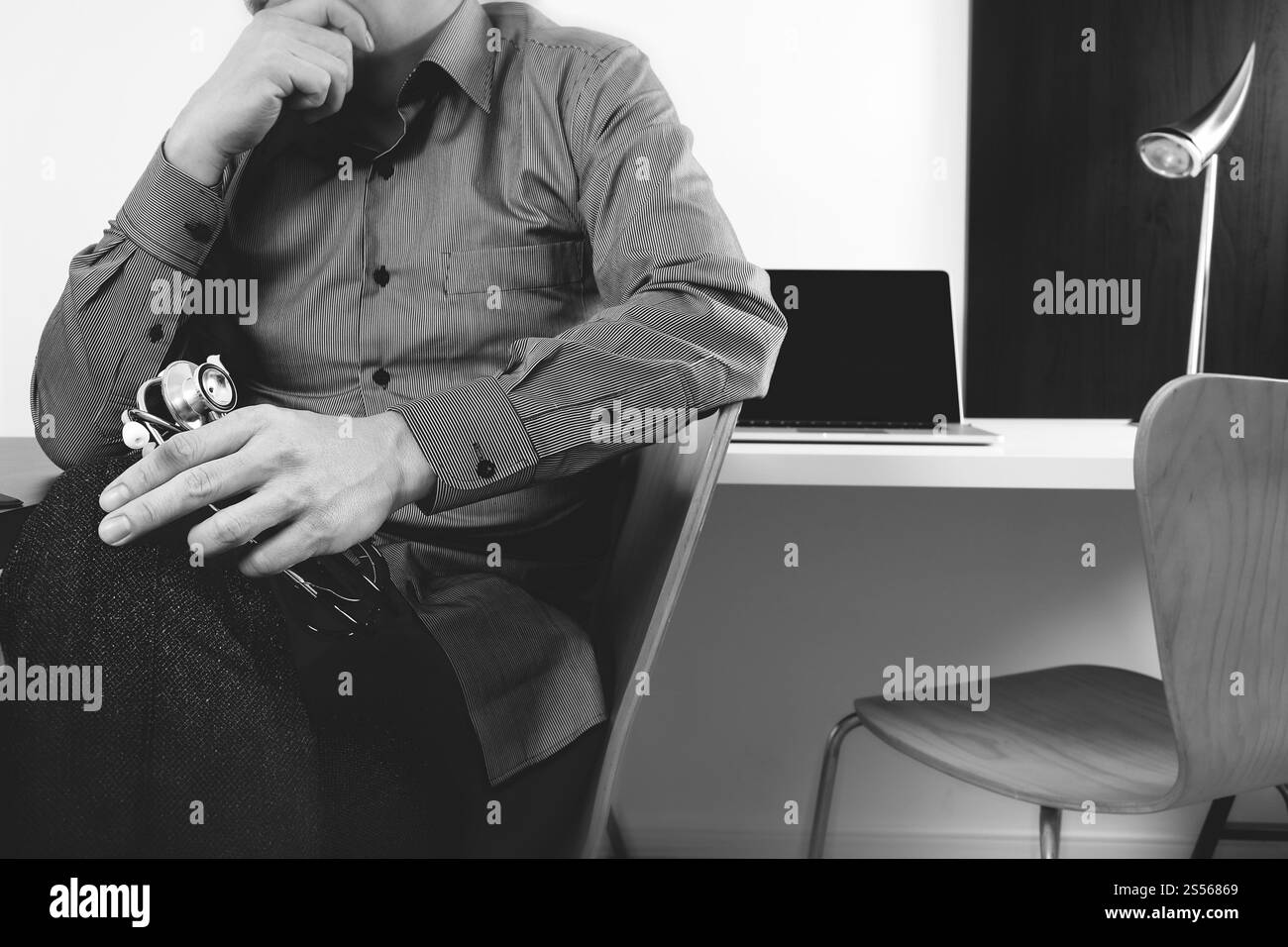 intelligente Arzt Stethoskop halten und denken mit Laptop-Computer auf dunklen Holz Schreibtisch im modernen Büro, schwarz / weiß Stockfoto