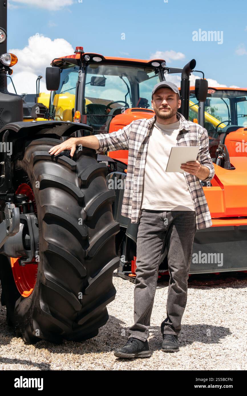 Kaukasischer Erwachsener mit digitalem Tablet in der Hand steht neben einem Bau- oder landwirtschaftlichen Traktor. Stockfoto