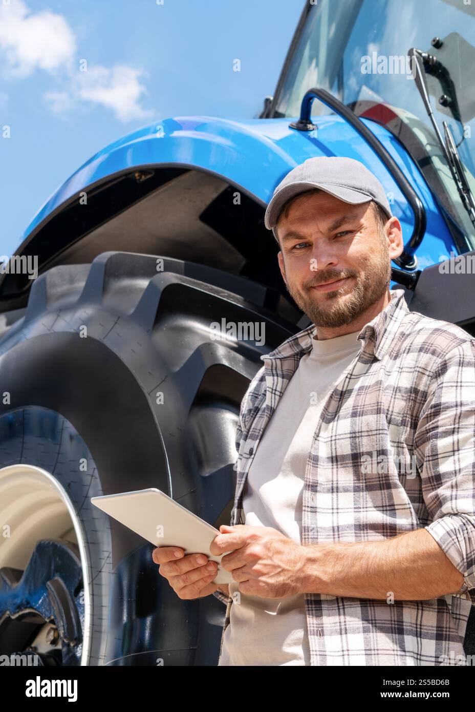 Vertikales Porträt eines fröhlichen männlichen Landarbeiters mit digitalem Tablet, das im Sommer in der Nähe eines blauen Traktors steht. Stockfoto