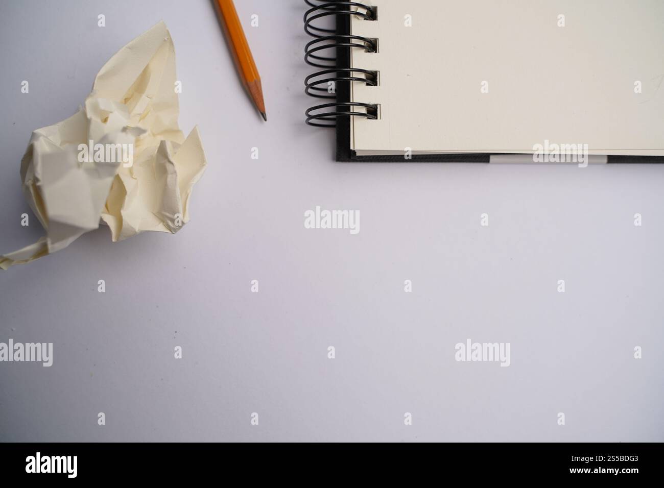 Minimalistischer Arbeitsbereich mit orangefarbenem Bleistift, zerknitterten Papieren und offenem Spiralnotizbuch auf weißem Schreibtisch. Perfekt für Themen wie Kreativität und Brainstom Stockfoto