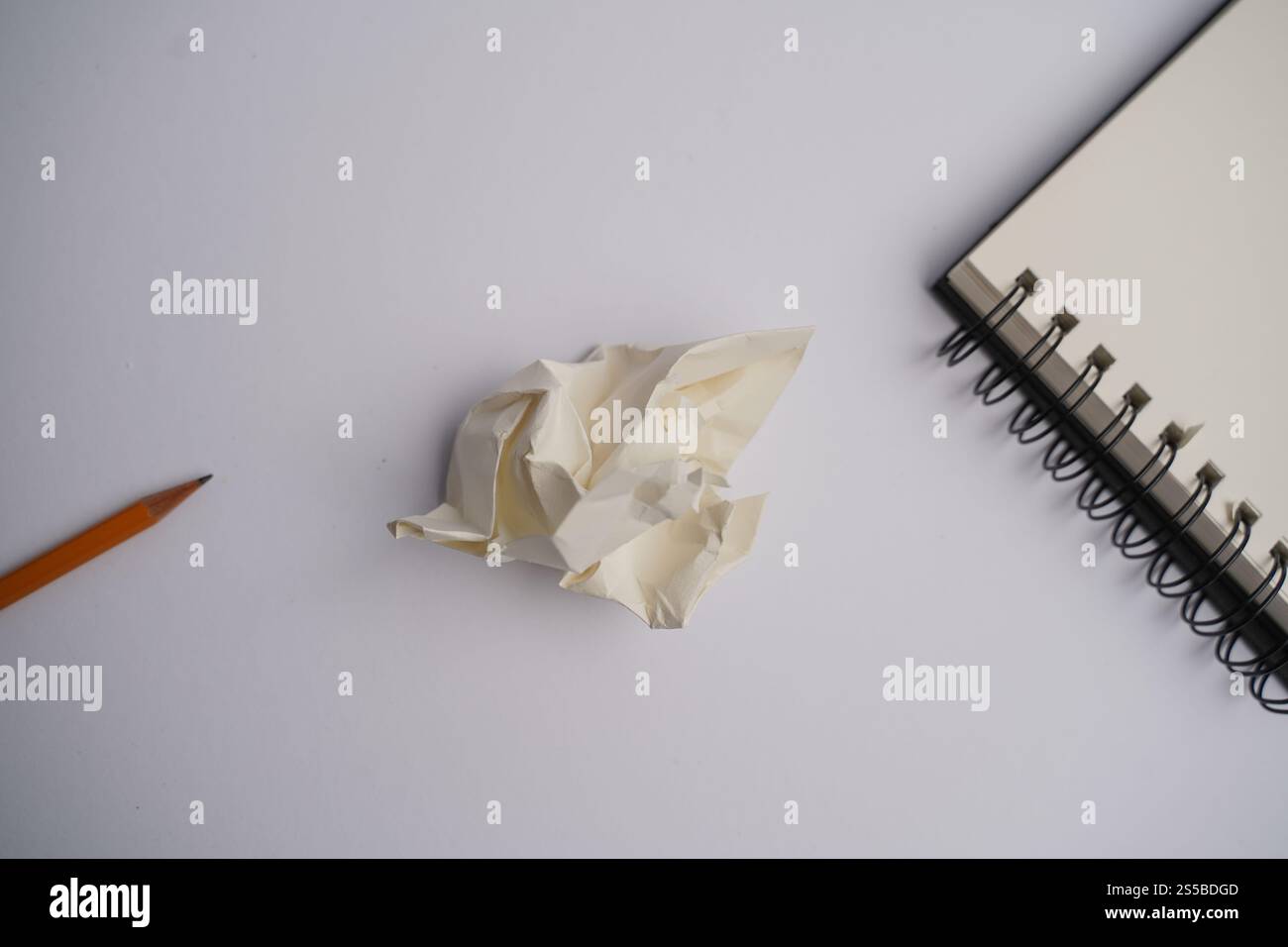Minimalistischer Arbeitsbereich mit orangefarbenem Bleistift, zerknitterten Papieren und offenem Spiralnotizbuch auf weißem Schreibtisch. Perfekt für Themen wie Kreativität und Brainstom Stockfoto