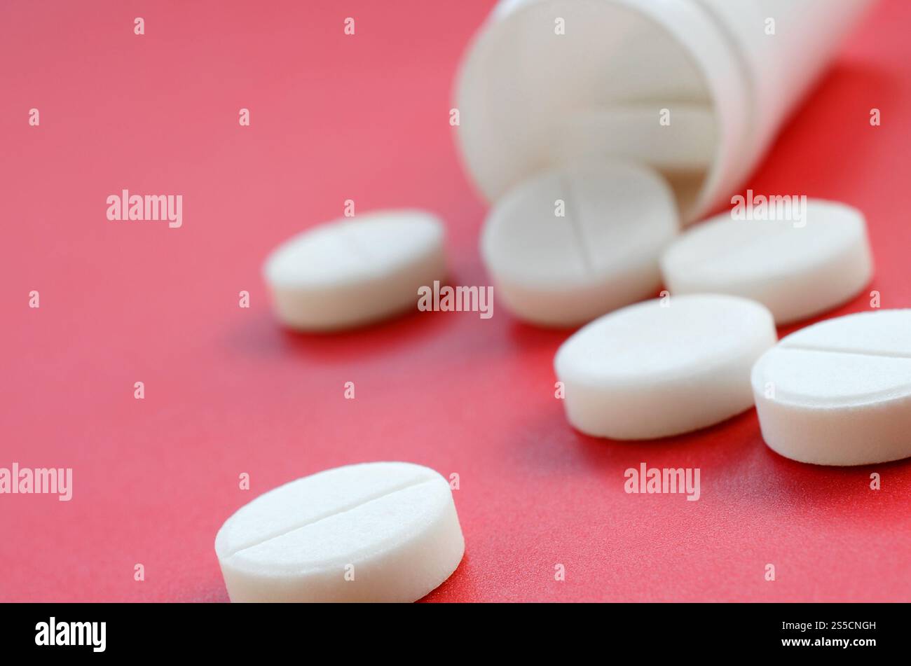Mehrere weiße Tabletten fallen auf der roten Oberfläche aus dem Kunststoffbehälter. Hintergrundbild zu medizinischen und pharmazeutischen Themen Stockfoto