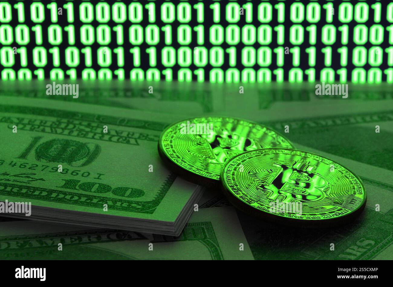 Zwei Bitcoins liegen auf einem Stapel von Dollarscheinen auf dem Hintergrund eines Monitors, der einen Binärcode aus hellgrünen Nullen und eine Einheit auf einem schwarzen zeigt Stockfoto