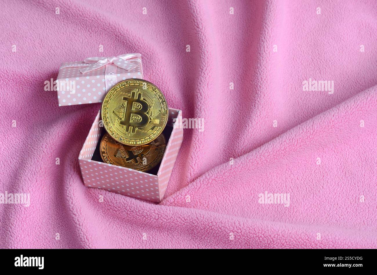 Der goldene Bitcoin liegt in einer kleinen rosafarbenen Geschenkbox mit einer kleinen Schleife auf einer Decke aus weichem und flauschigem hellrosa Fleecestoff mit einer großen Anzahl Stockfoto
