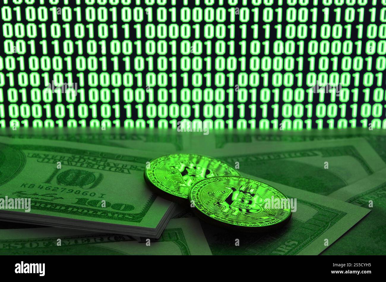 Zwei Bitcoins liegen auf einem Stapel von Dollarscheinen auf dem Hintergrund eines Monitors, der einen Binärcode aus hellgrünen Nullen und eine Einheit auf einem schwarzen zeigt Stockfoto