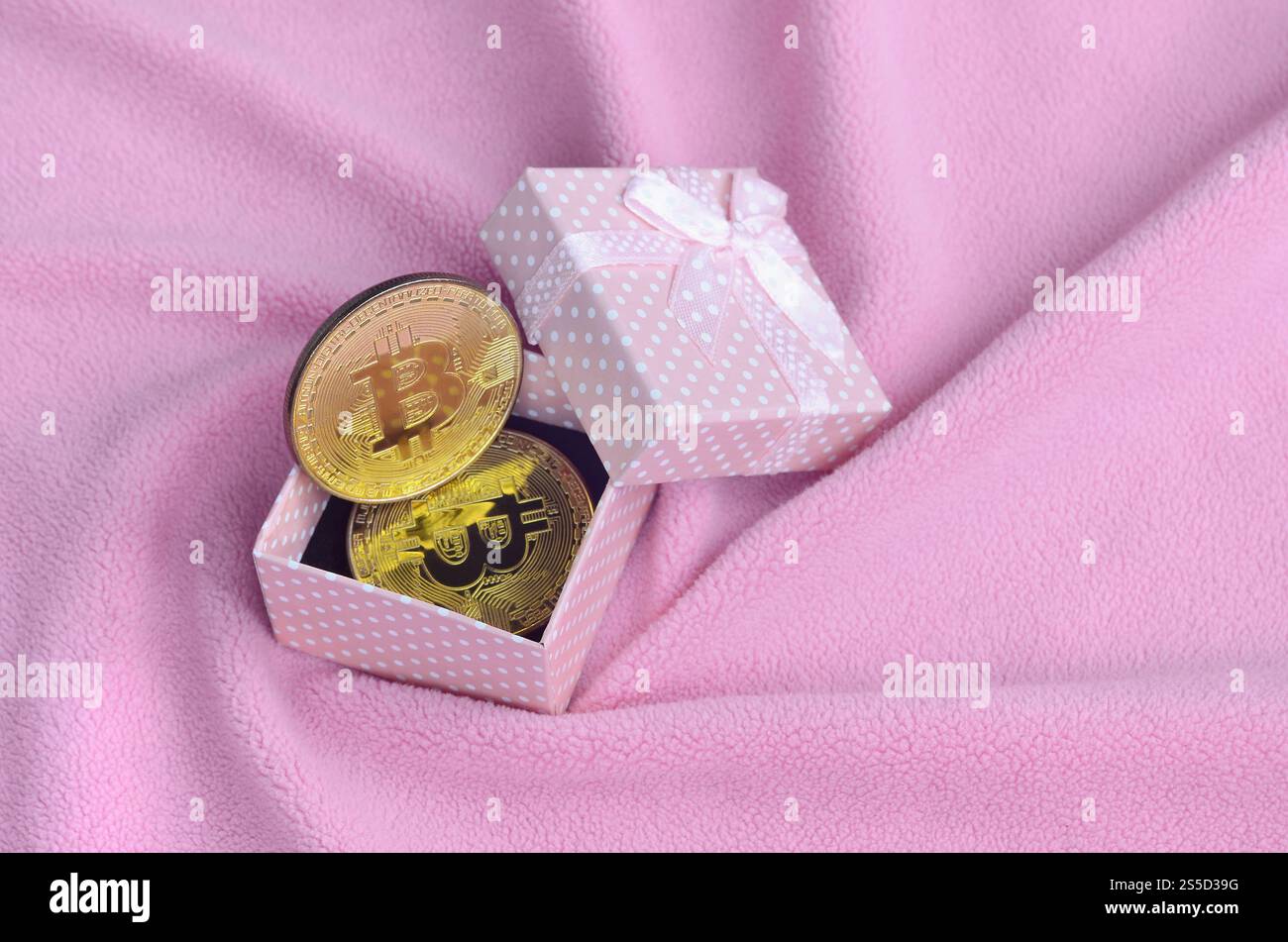 Der goldene Bitcoin liegt in einer kleinen rosafarbenen Geschenkbox mit einer kleinen Schleife auf einer Decke aus weichem und flauschigem hellrosa Fleecestoff mit einer großen Anzahl Stockfoto