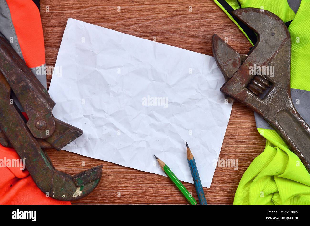 Ein zerknittertes Blatt Papier mit zwei Bleistiften, umgeben von grünen und orangen Arbeitsuniformen und verstellbaren Schraubenschlüsseln. Stillleben in Verbindung mit Stockfoto