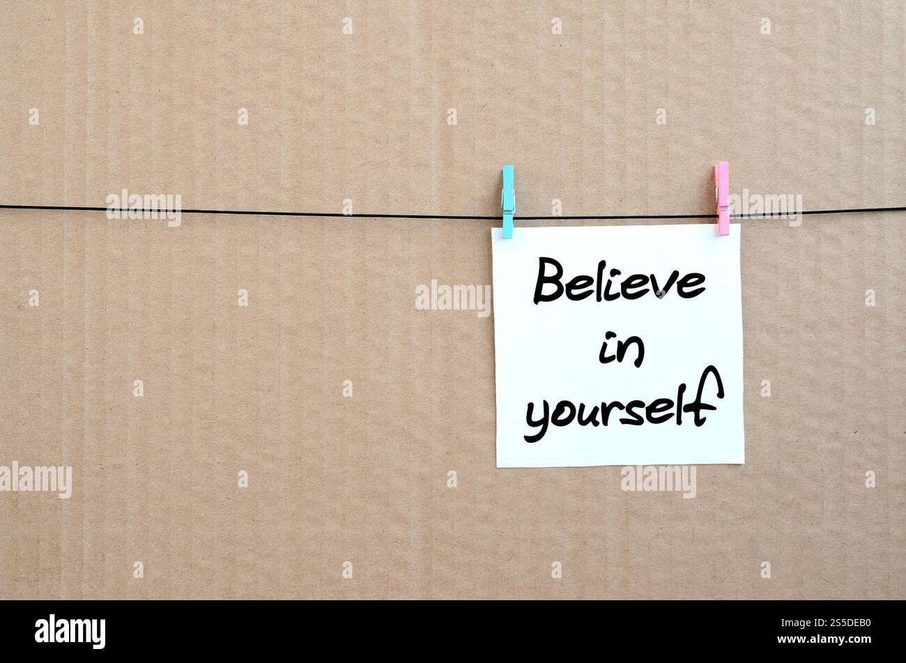 Glauben Sie an sich selbst. Die Notiz ist auf einem weißen Aufkleber geschrieben, der mit einem Kleiderschrank an einem Seil auf einem Hintergrund aus braunem Karton hängt Stockfoto Glauben Sie an sich selbst. Die Notiz ist auf einem weißen Aufkleber geschrieben, der mit einem Kleiderschrank an einem Seil auf einem Hintergrund aus braunem Karton hängt Stockfoto
