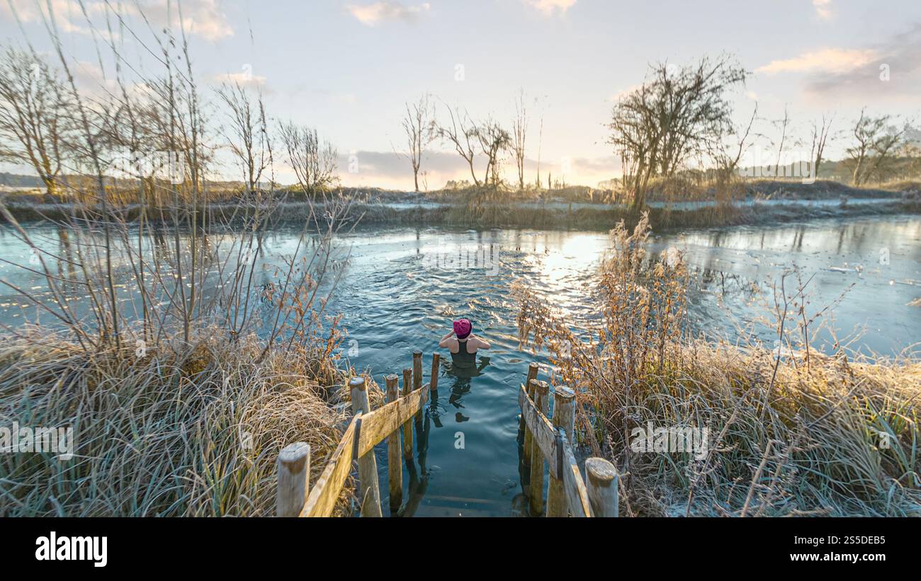 Weibliche Schwimmerin, von hinten gesehen, mit Wollmütze, in der Nähe der Holzstufen eines gefrorenen Schwimmteichs. York, Großbritannien Stockfoto