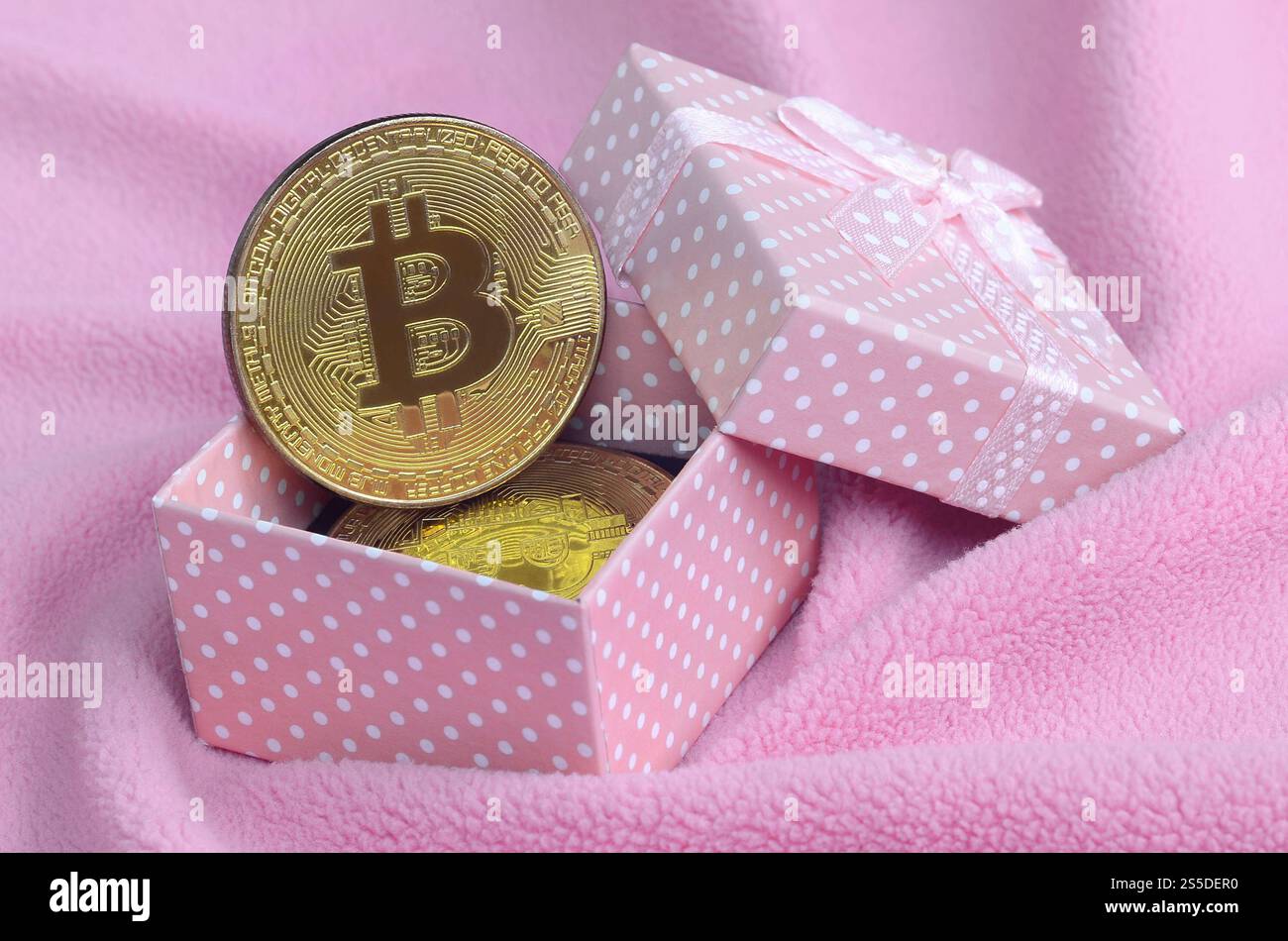Der goldene Bitcoin liegt in einer kleinen rosafarbenen Geschenkbox mit einer kleinen Schleife auf einer Decke aus weichem und flauschigem hellrosa Fleecestoff mit einer großen Anzahl Stockfoto