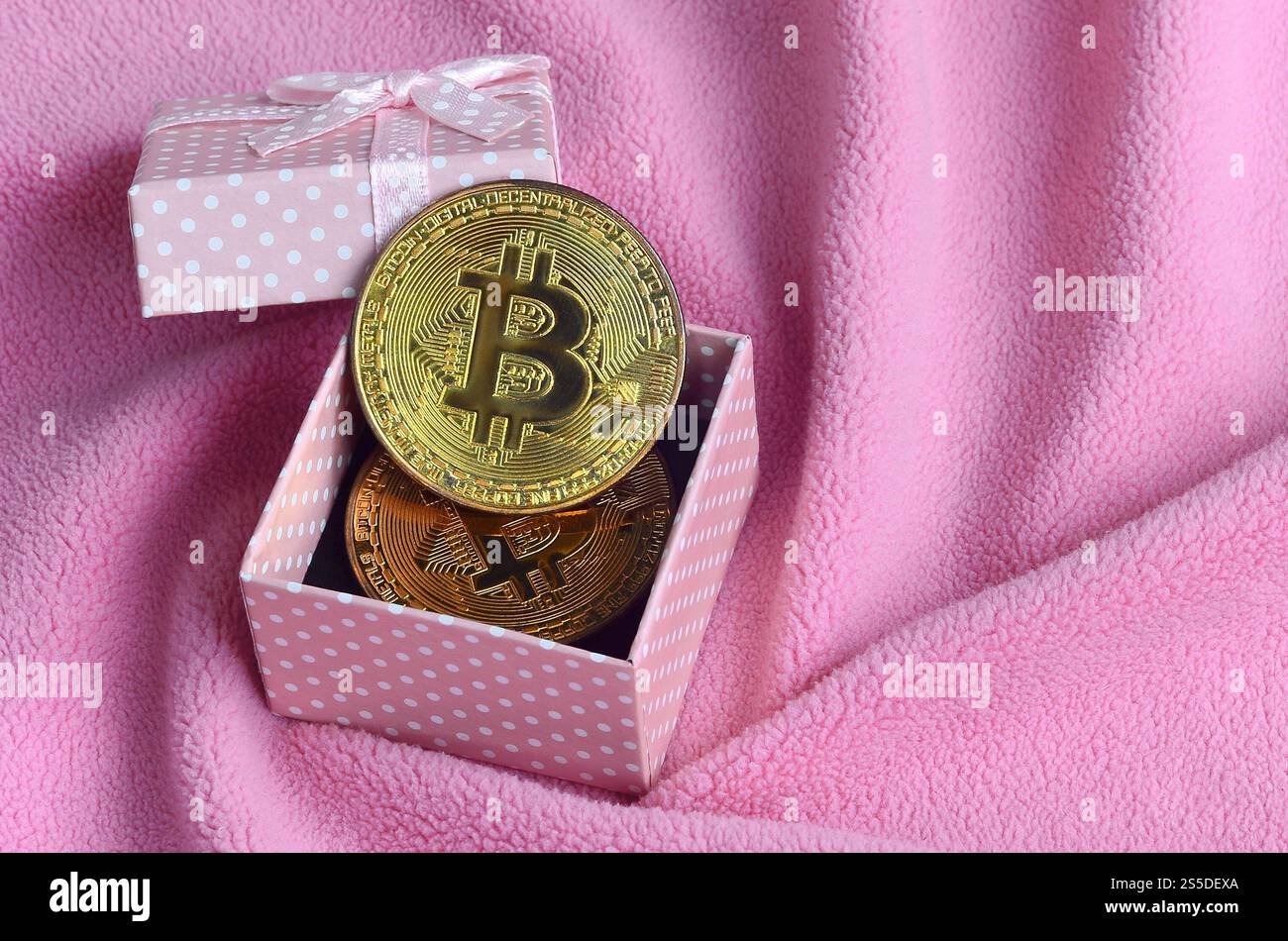 Der goldene Bitcoin liegt in einer kleinen rosafarbenen Geschenkbox mit einer kleinen Schleife auf einer Decke aus weichem und flauschigem hellrosa Fleecestoff mit einer großen Anzahl Stockfoto