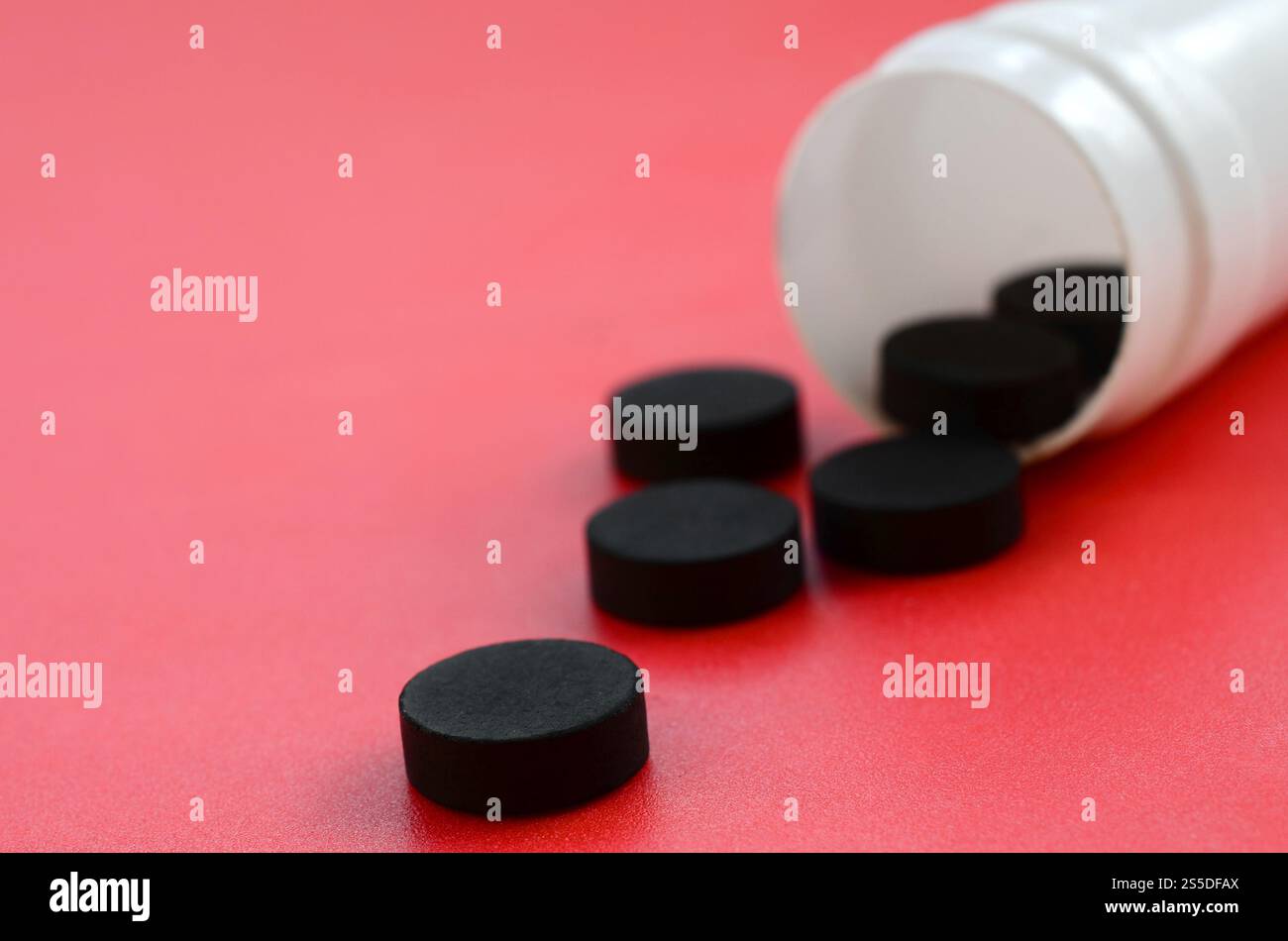 Mehrere schwarze Tabletten fallen aus dem Kunststoffbehälter auf der roten Oberfläche. Hintergrundbild zu medizinischen und pharmazeutischen Themen. Aktivkohle Stockfoto