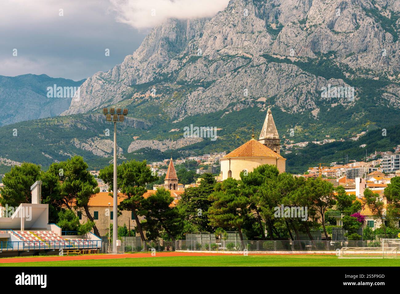 Outdoor-Sportfußballstadion mit Laufbahnen, Sitzplätzen und Flutlichtern, mit der historischen Stadt Makarska und zerklüfteten Bergen im Hintergrund, Kroatien Stockfoto