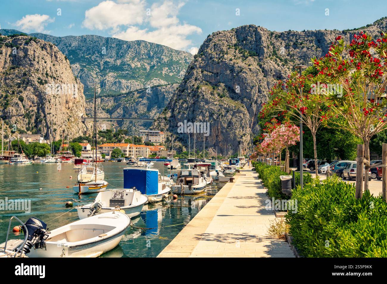 Idyllische Küstenszene mit Booten, die entlang einer von Bäumen gesäumten Promenade vor dem Hintergrund majestätischer Berge und eines klaren blauen Himmels in Omis, Kroatien, vertäut sind Stockfoto
