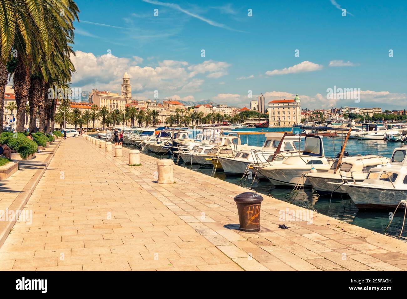 Sonniger Tag an einem mediterranen Yachthafen mit verankerten Booten und einer von Bäumen gesäumten Promenade, die zu historischen Gebäuden in Split, Kroatien, führt Stockfoto