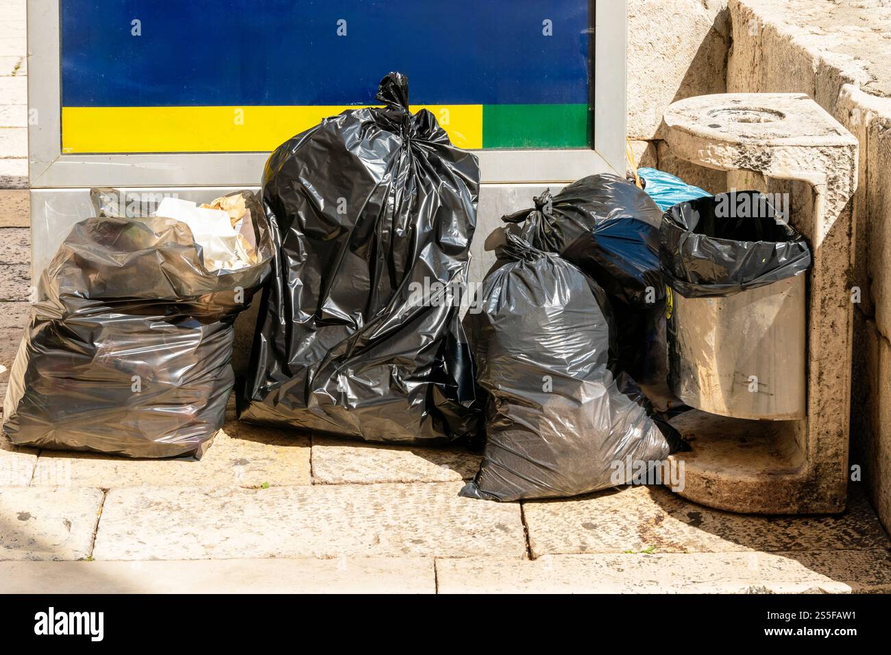 Schwarze Müllsäcke stapelten sich an einem sonnigen Tag neben einem Steinmüllbehälter mit einem Informationsschild im Hintergrund, Split, Kroatien Stockfoto