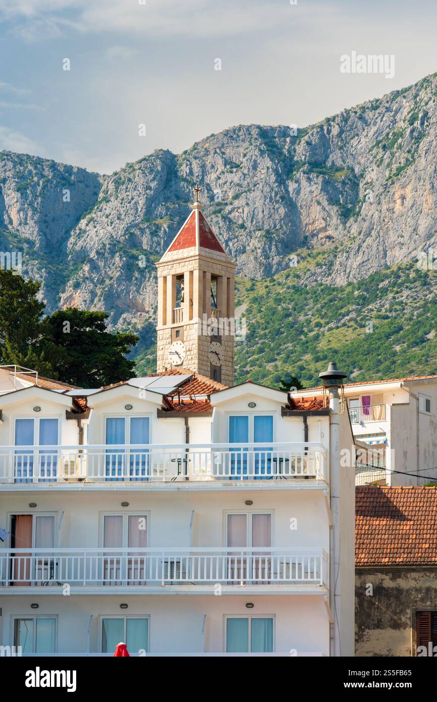 Weißes Gebäude mit einem roten Dach und einem Kirchturm vor einer Bergkulisse unter einem klaren blauen Himmel, Drvenik, Kroatien. Stockfoto