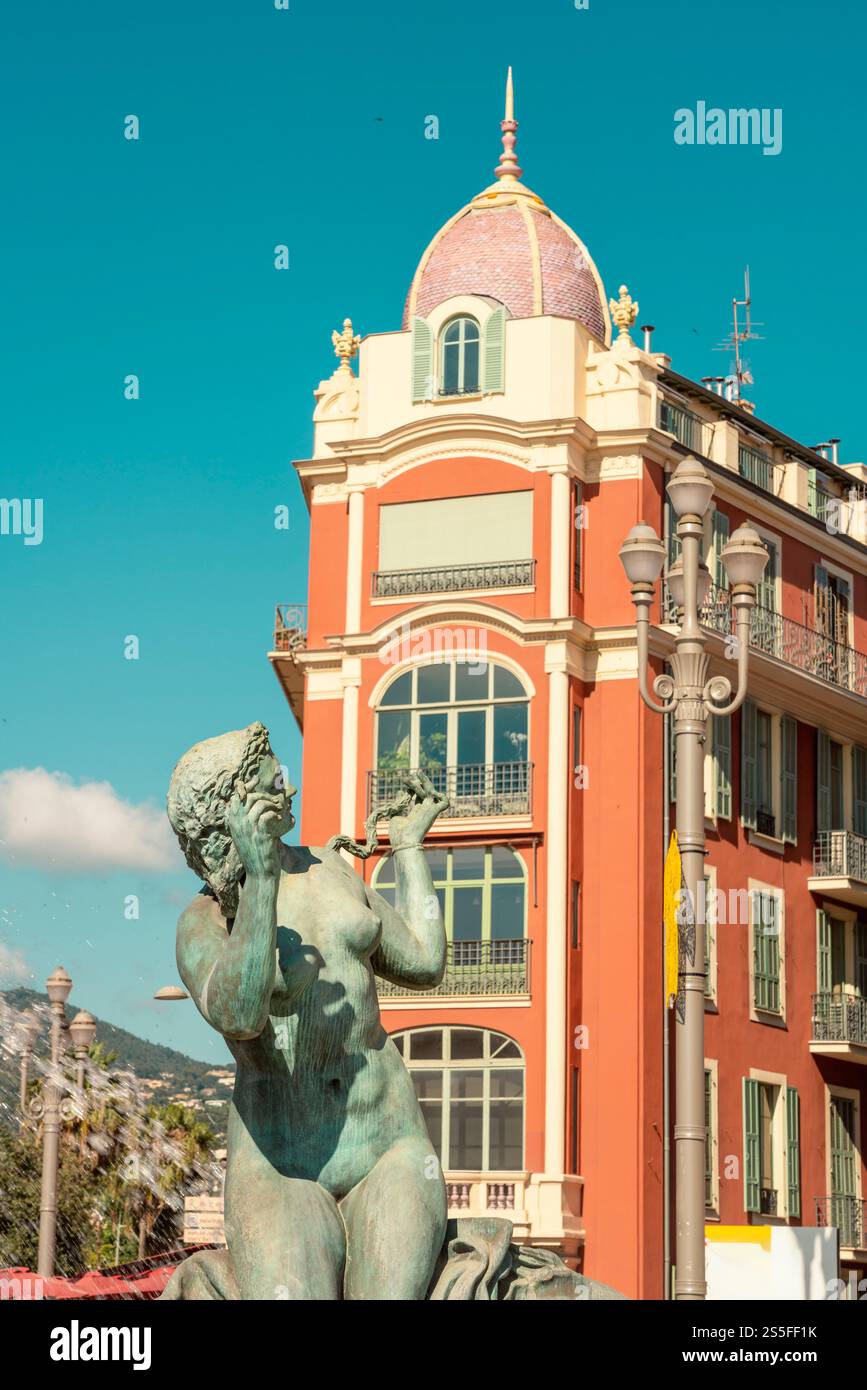 Eine Bronzestatue vor einem lebhaften orangen Gebäude mit einer bunten gekachelten Kuppel unter einem klaren blauen Himmel von Fontaine du Soleil am Place Massena, Nizza, Frankreich Stockfoto