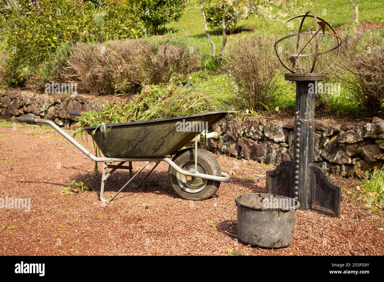 Eine Schubkarre gefüllt mit Grasschnitt neben einer Metallsonnenuhr und einem Eimer in einem Garten in Auckland, Neuseeland Stockfoto