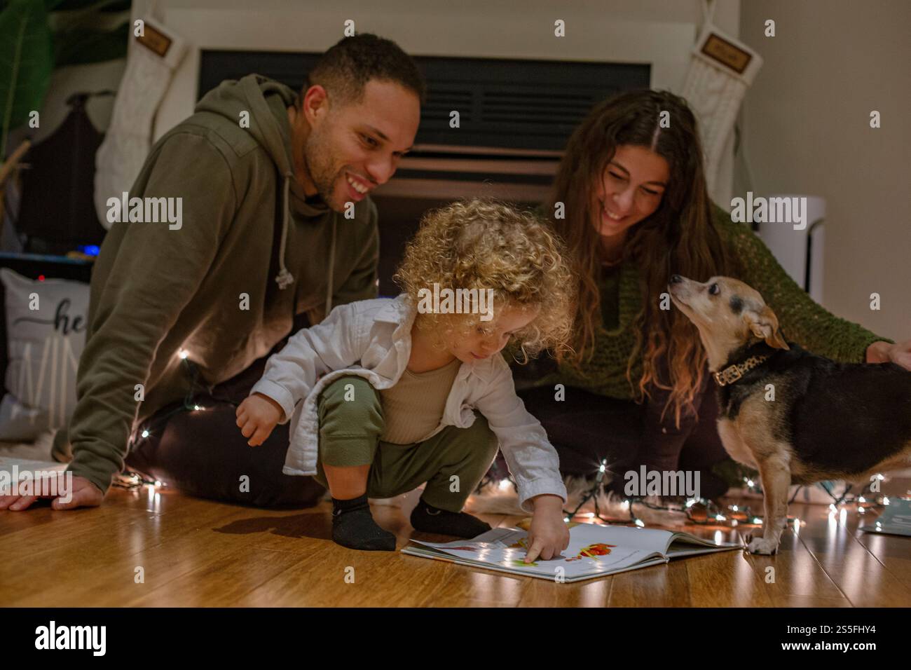 Eine Familie genießt die gemeinsame Zeit, während ein Kleinkind mit lockigen Haaren ein Buch mit leuchtenden Lichtern liest, begleitet von einem aufmerksamen Hund, Ohio, USA Stockfoto