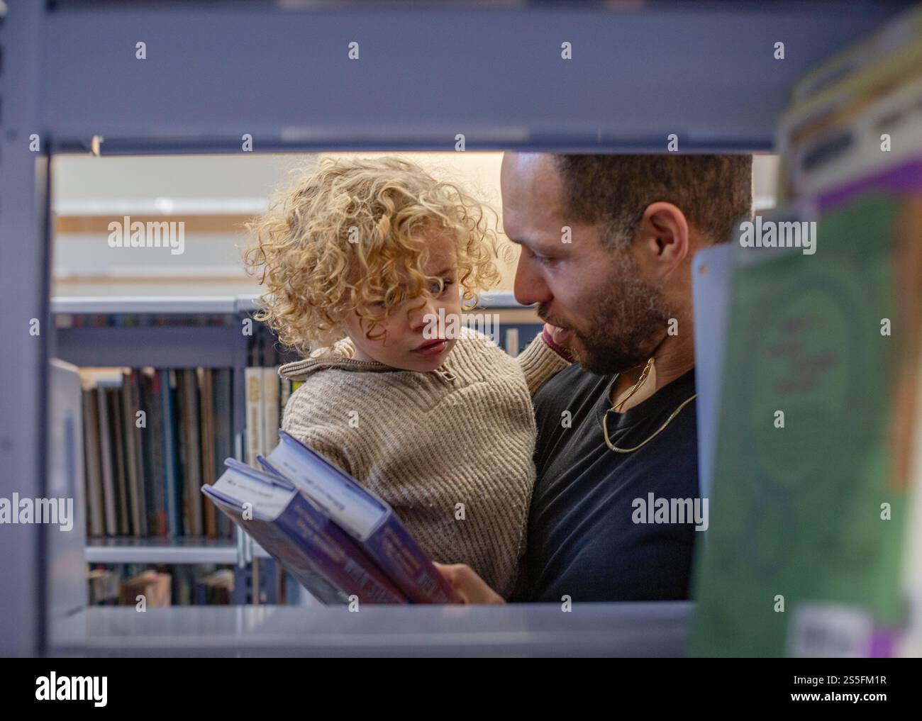 Lockhaariges Kind schaut neugierig auf ein Buch, das von einem bärtigen Erwachsenen in einer Bibliothek gehalten wird. Cleveland, Ohio, USA Stockfoto