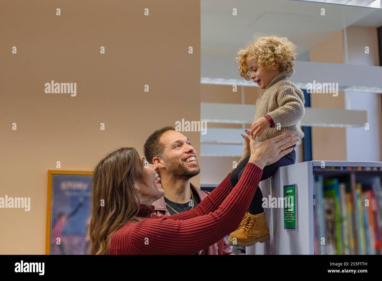 Lächelnde Familie spielt mit einem Kleinkind, das von seinen Eltern in einem Bibliotheksraum mit Bücherregalen hochgehoben wird. Cleveland, Ohio, USA Stockfoto