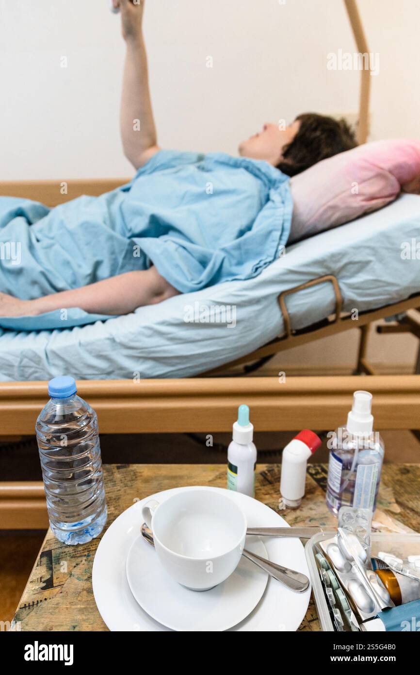 Weißer Becher mit Untertasse und Medikamenten auf dem Tisch und kranke Frau hält Griff des Homecare Krankenhausbetts auf dem Hintergrund zu Hause Stockfoto