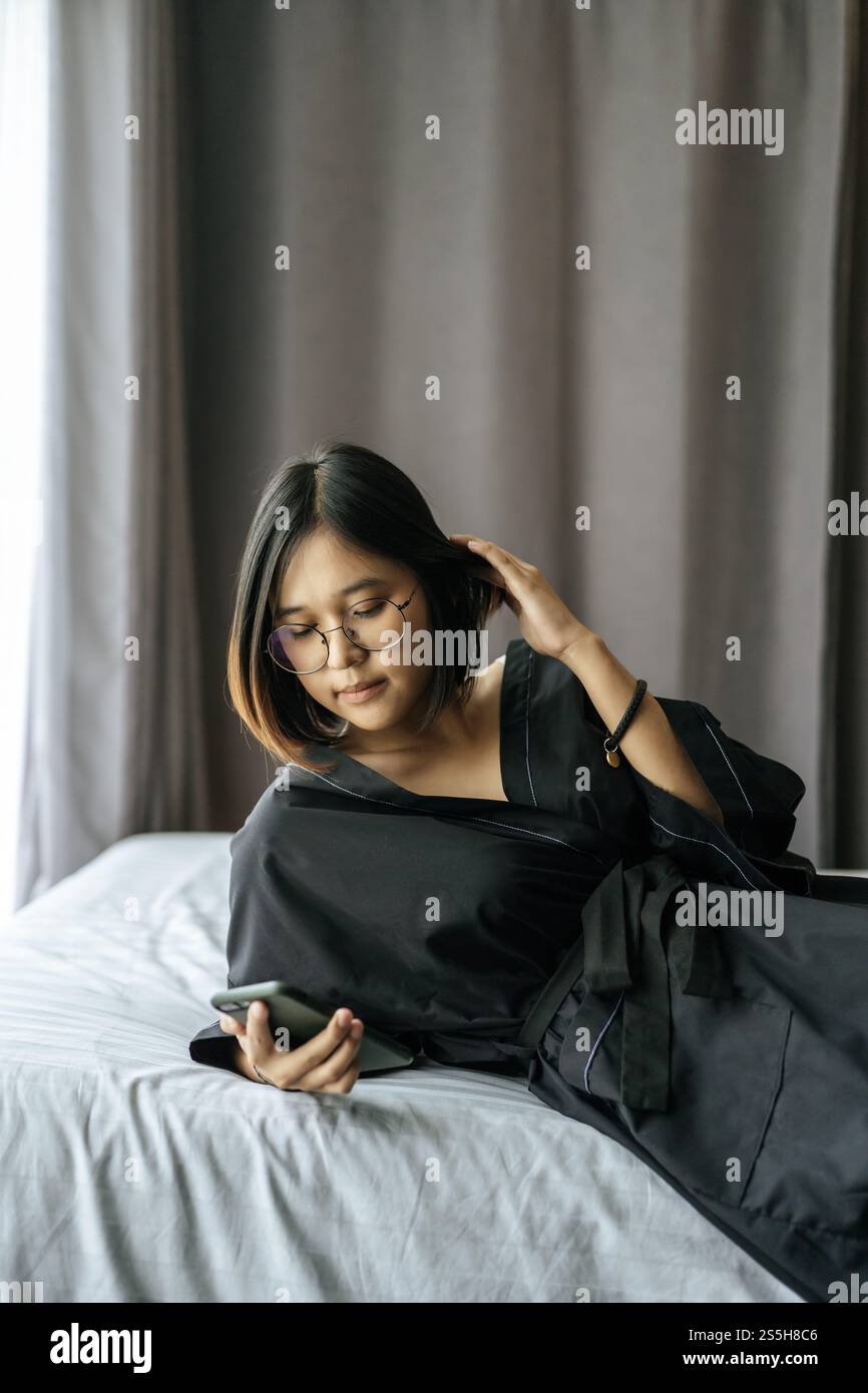 Frau mit schwarzem Hemd, die auf dem Bett liegt und Smartphone spielt. Stockfoto