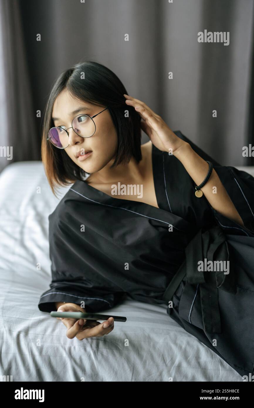 Frau mit schwarzem Hemd, die auf dem Bett liegt und Smartphone spielt. Stockfoto