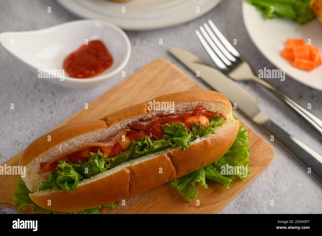 Selektiver Fokus Hotdog und Speck in Brot mit Salat auf Holzbrett, verschwommener Tomatensauce, Gabel und Messer, Kopierraum Stockfoto