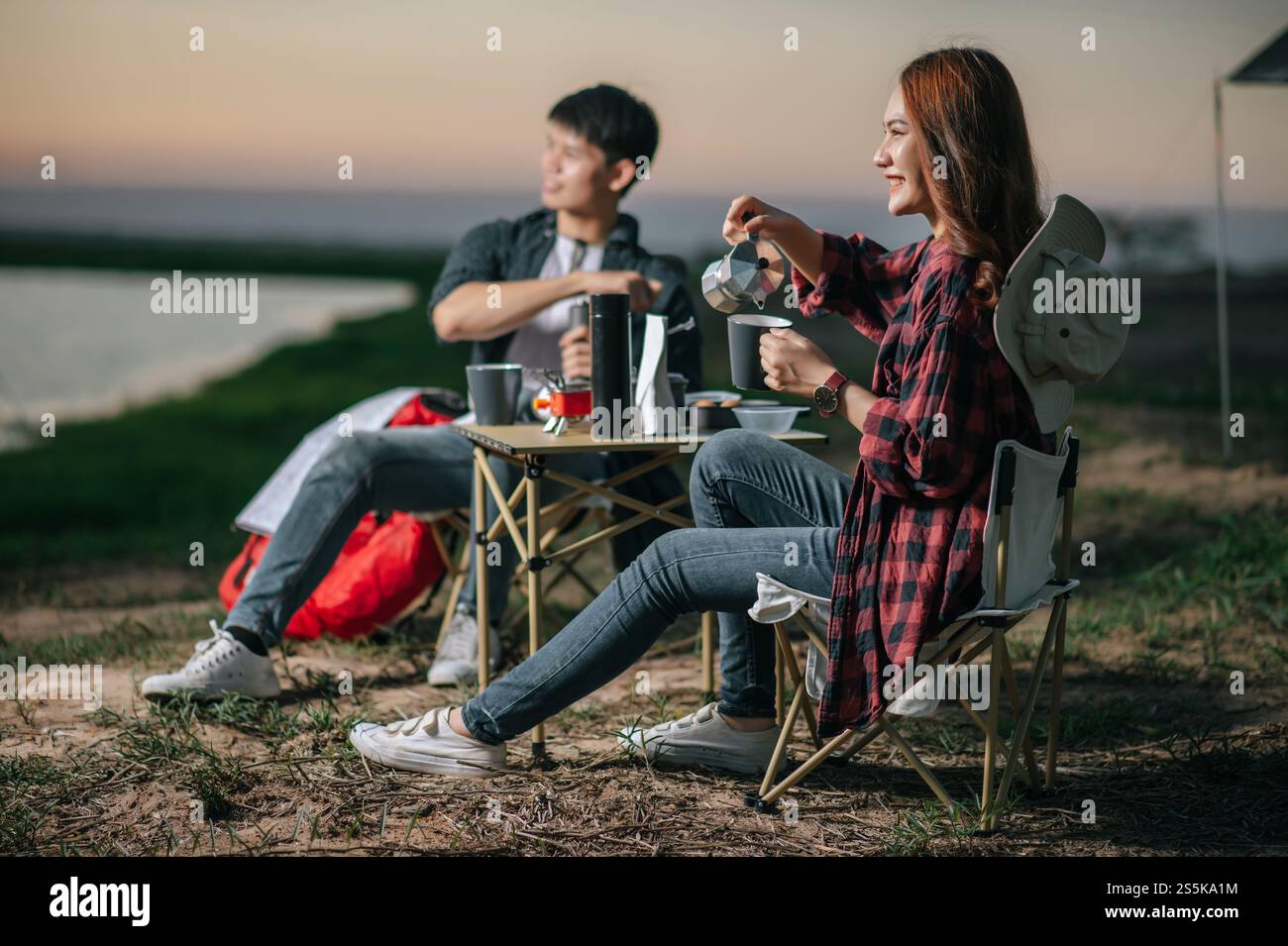 Fröhliches junges Backpacker-Paar, das vor dem Zelt im Wald mit Kaffee-Set sitzt und während eines Campingausflugs im Sommer frische Kaffeemühle zubereitete Stockfoto