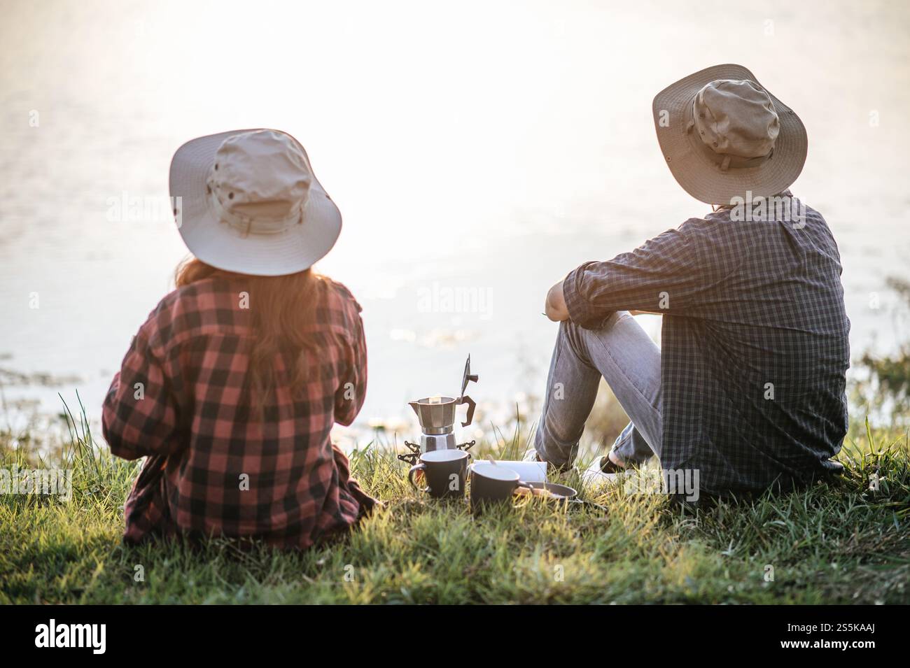 Fröhliches junges Backpacker-Paar, das auf Gras sitzt und sich am frühen Morgen über den See freut und während des Campingausflugs frische Kaffeemühle zubereitete Stockfoto