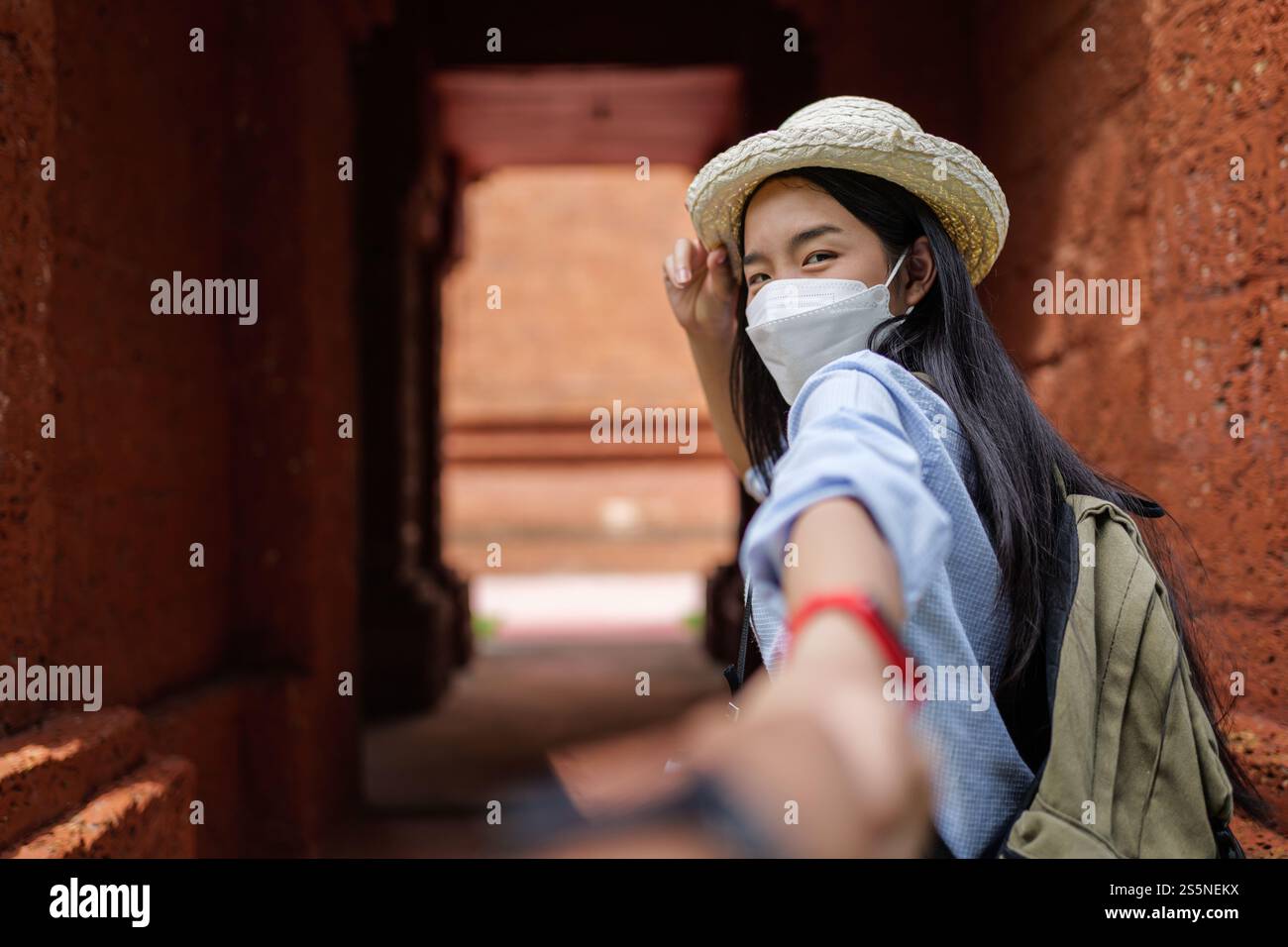 Asiatische junge Frau, die Schutzmaske trägt und die Hand einer ernten Person hält, während sie geht, verschwommener Hintergrund mit der antiken Stätte, neue normale des Reisenden Stockfoto
