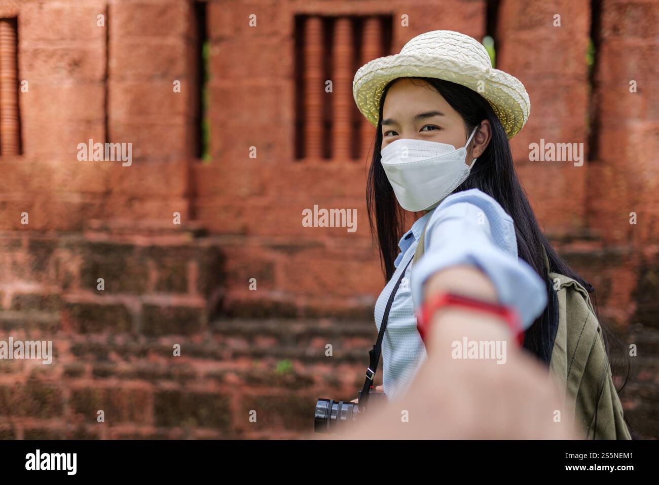 Asiatische junge Frau, die Schutzmaske trägt und die Hand einer ernten Person hält, während sie geht, verschwommener Hintergrund mit der antiken Stätte, neue normale des Reisenden Stockfoto