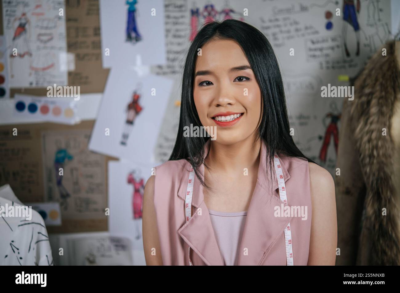 Portrait junge Designerin, die mit ihren Modekleidern am Kleiderbügel steht, Maßband am Hals, Kleiderskizze an Bord, Verstecken Sie sie und schauen Sie nach Stockfoto