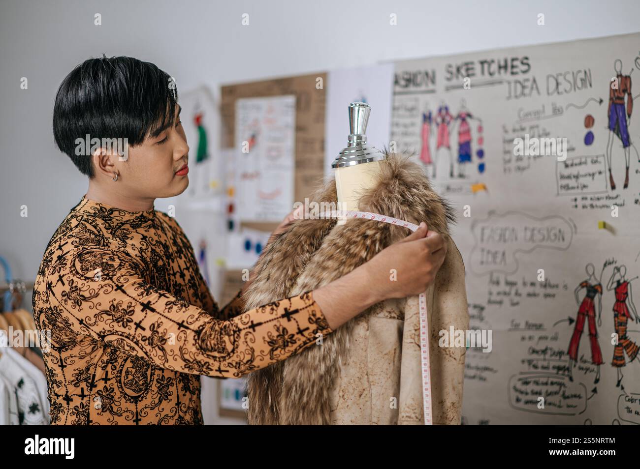 Porträt eines jungen asiatischen Schneiders, der an neuem Bekleidungsdesign am Tisch im Modestudio misst. Designer und LGBTQ-Konzept. Stockfoto