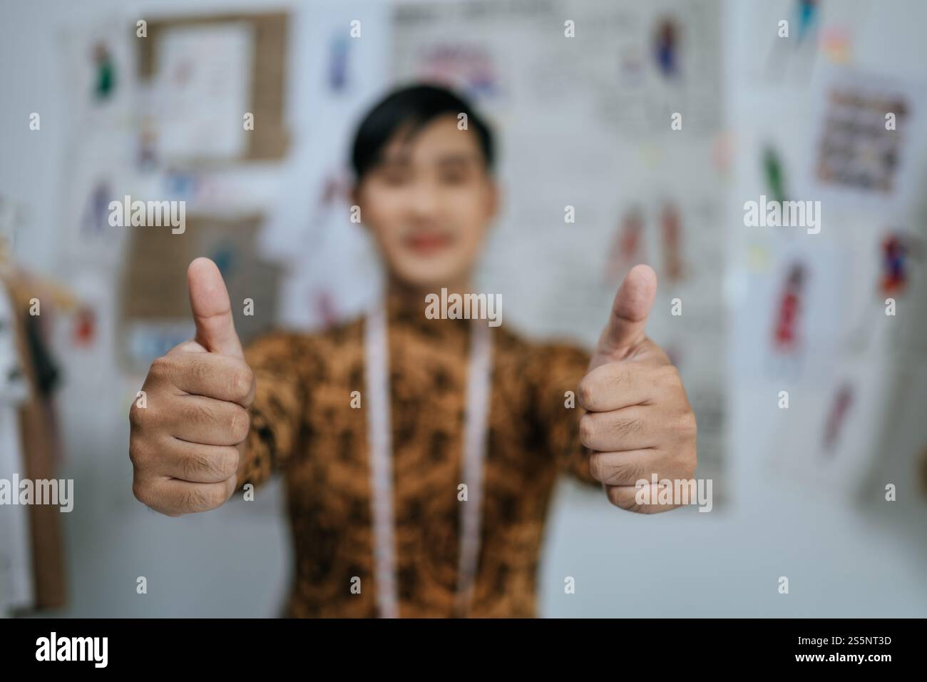 Fokussierter Finger des professionellen asiatischen jungen Schneiders mit Maßband am Hals Daumen nach oben und Blick auf die Kamera mit Zeichnungen Hintergrund im Studio. Stockfoto
