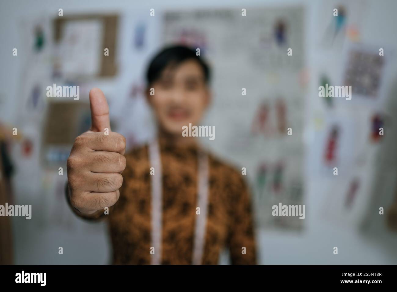 Fokussierter Finger des professionellen asiatischen jungen Schneiders mit Maßband am Hals Daumen nach oben und Blick auf die Kamera mit Zeichnungen Hintergrund im Studio. Stockfoto
