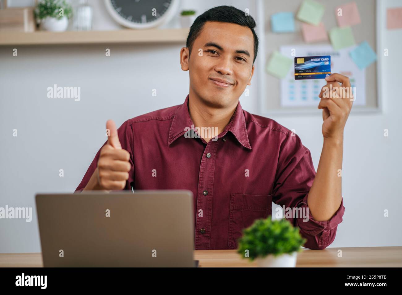 Mann, der am Laptop arbeitet, Daumen hoch und Kreditkarte zeigt Stockfoto